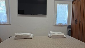 Habitación de hotel con una cama grande, dos ventanas con cortinas, un televisor de pantalla plana montado en la pared y un armario de madera.