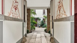 A corridor with a tiled floor leading to a courtyard with plants and a building in the background. - Isola Stay - A beautiful little nest in Isola, in the heart of Milan (Milano)