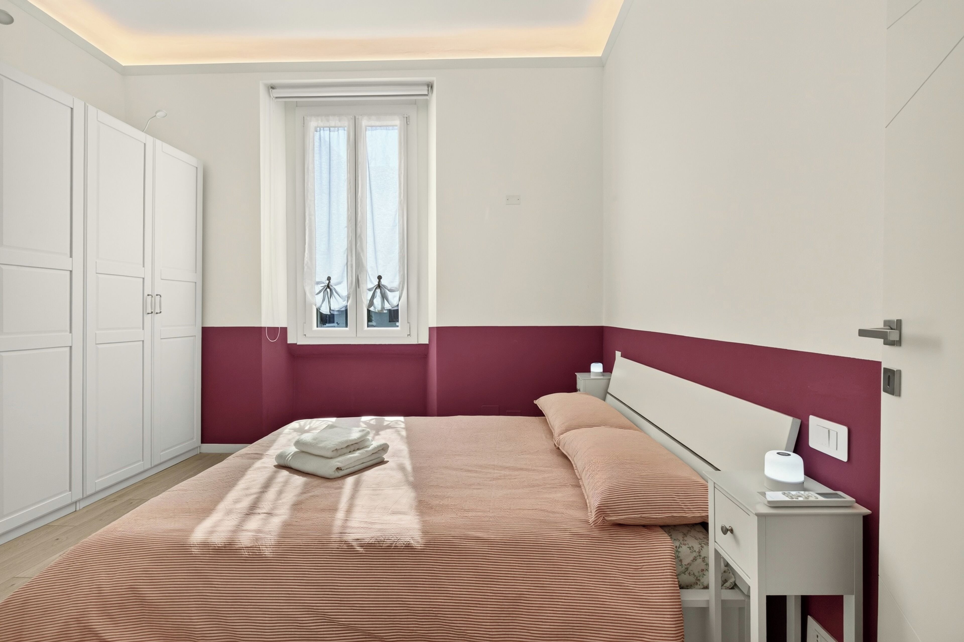 A bedroom with a bed, a window with curtains, a bedside table with a lamp, and a wardrobe.