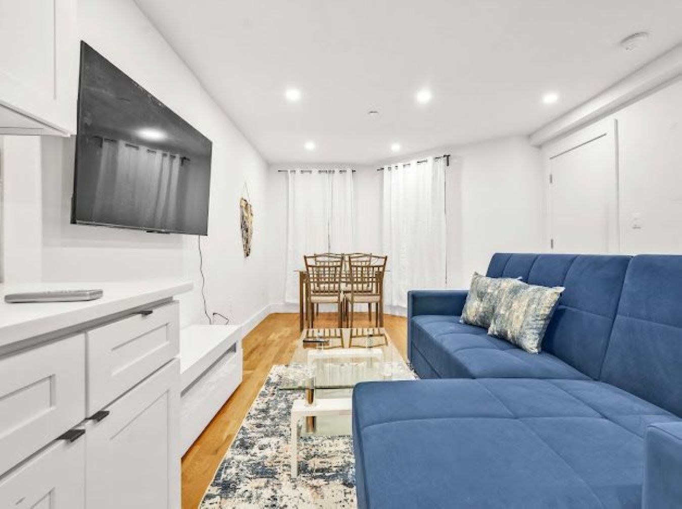 A modern living room with a blue sofa, a flat-screen TV mounted on the wall, a wooden dining table, and a white cabinet.