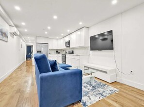 A modern living room with a blue sofa, a flat-screen TV mounted on the wall, and a kitchen in the background.
