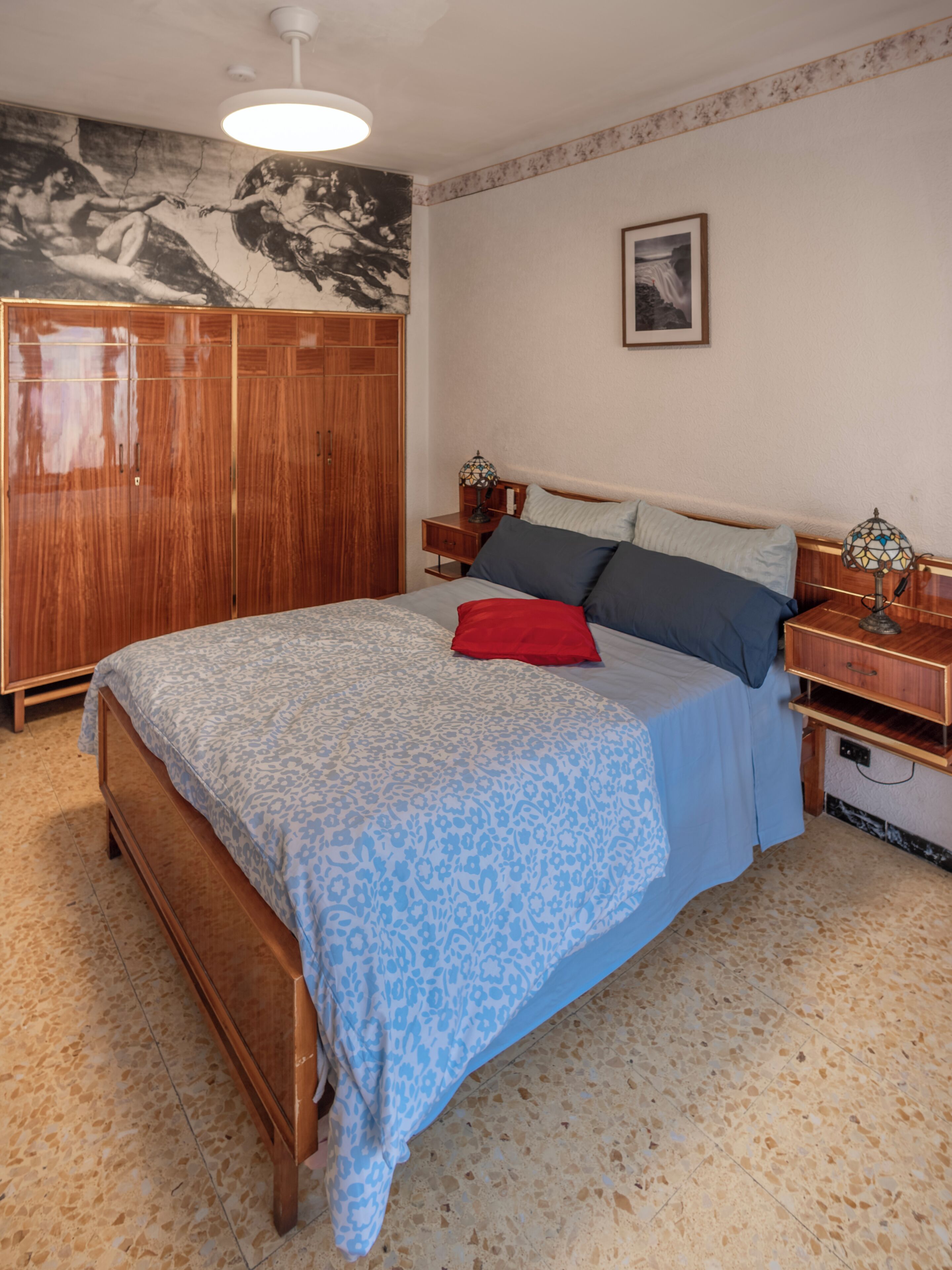 Un dormitorio con una cama, armario de madera, mesitas de noche, una lámpara y un cuadro enmarcado en la pared.