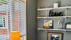 Una stanza con una finestra, una lampada, uno scaffale con oggetti decorativi e un quadro incorniciato.