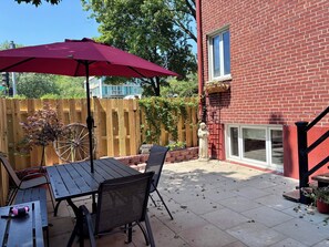 Eine Terrasse mit Tisch und Stühlen, einem roten Sonnenschirm, einem Holzzaun und einem Backsteinhaus mit Fenster.