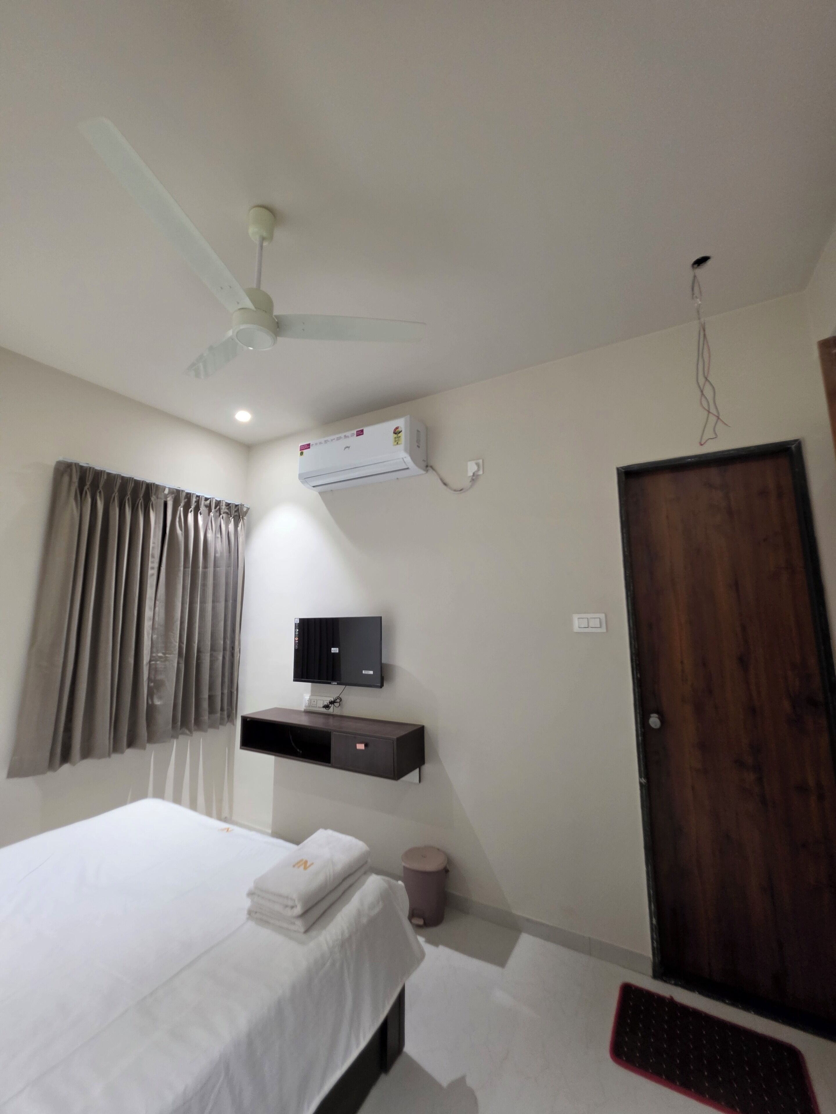 Habitación de hotel con cama, aire acondicionado de pared, televisor, puerta de madera y ventilador de techo.