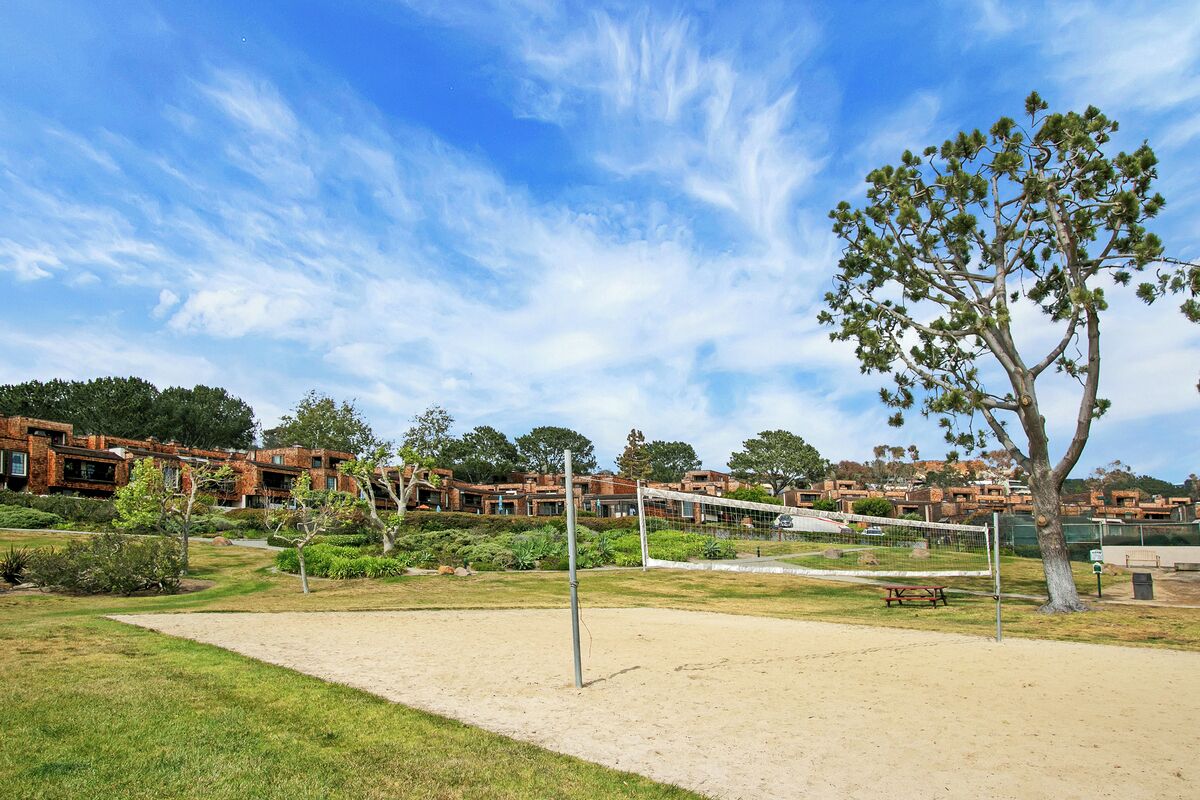 A sandy volleyball court with a net, surrounded by a landscaped area with trees and shrubs, and brick buildings in the background.