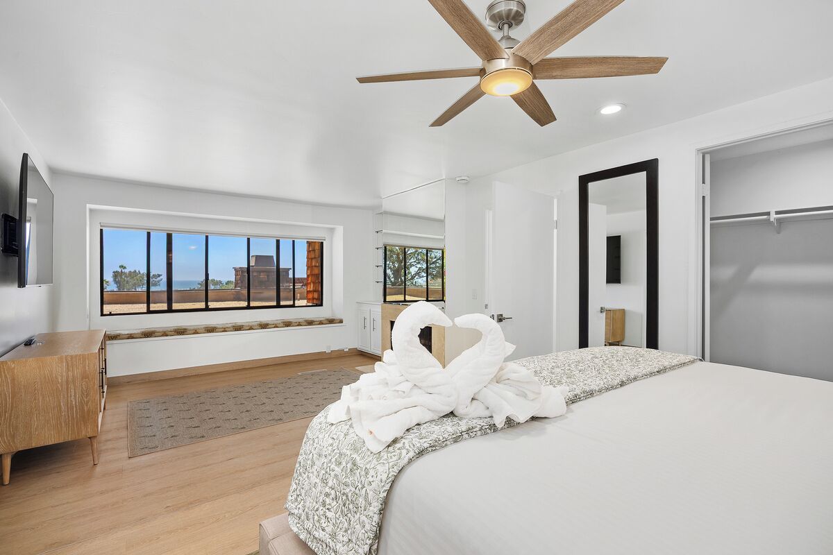 A hotel room with a bed, a ceiling fan, a television, a window with a view, and a wooden dresser.