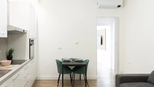 A compact kitchen with white cabinets, a dining table with green chairs, and a grey sofa in a bright room with wooden flooring. - IMB23 • Central Apartment in Milan • (Milano)