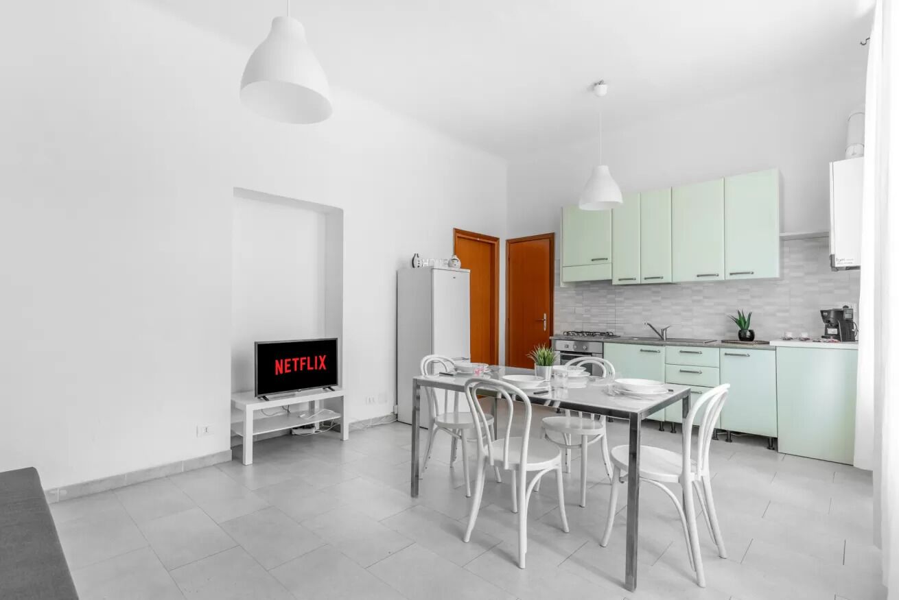 Une cuisine moderne avec des meubles blancs, un coin repas avec des chaises blanches, un téléviseur diffusant Netflix et des luminaires suspendus.