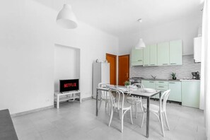 A modern kitchen with white cabinets, a dining area with white chairs, a TV displaying Netflix, and pendant lighting. - VE2 [Island] Cool Apartment • Exclusive • (Milano)
