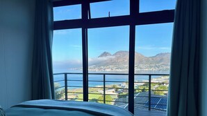 Quarto com uma janela ampla, uma cama e vista para uma paisagem costeira com montanhas.