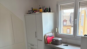 Ett kompakt kök med vita skåp, en bänkskiva, ett fönster med persienner och utsikt över byggnaderna utanför.