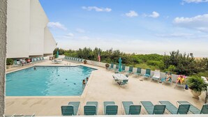 A swimming pool with lounge chairs and umbrellas, surrounded by greenery and a view of the beach. - Includes Xplorie Activities & Linens. PYRAMID-1BR OF condo w/outdoor pool. (Ocean City)