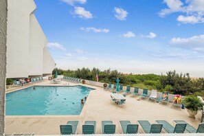 A swimming pool with lounge chairs and umbrellas, surrounded by greenery and a view of the beach. - Includes Xplorie Activities & Linens. PYRAMID-1BR OF condo w/outdoor pool. (Ocean City)