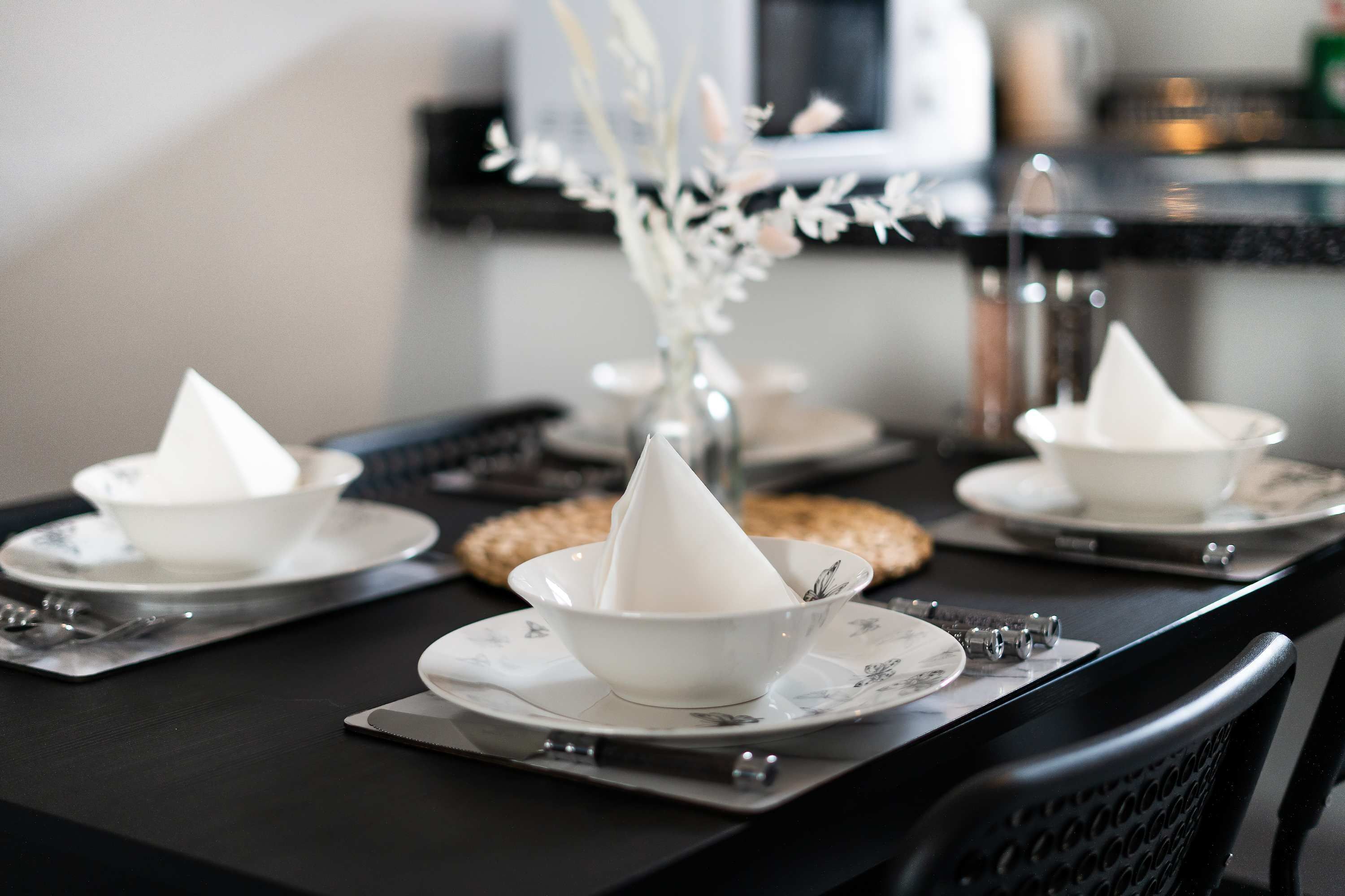 A dining table with white bowls and plates, a vase with white flowers, and a microwave in the background.
