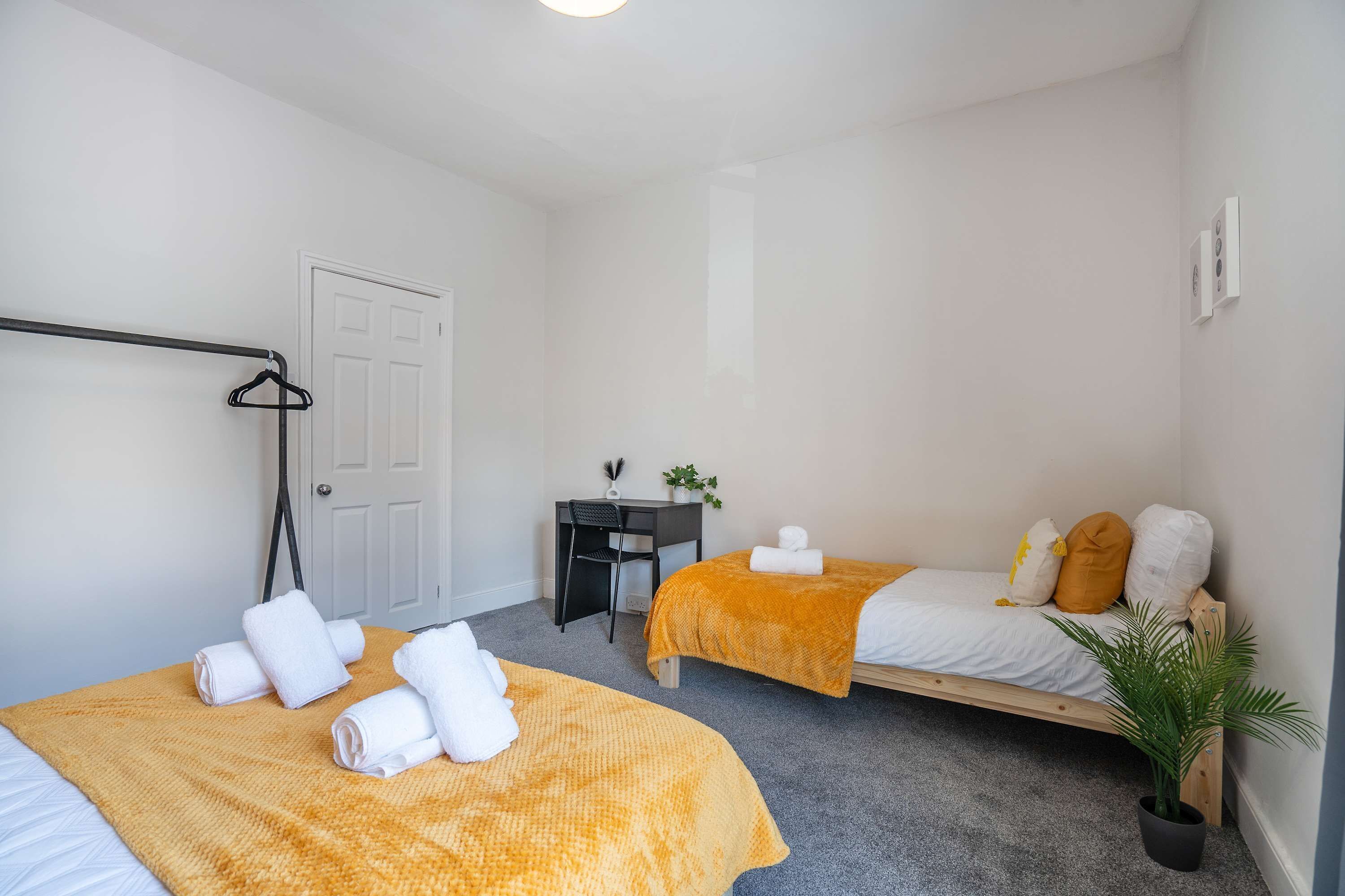 A room with two beds, a desk, a chair, a potted plant, and a coat rack.