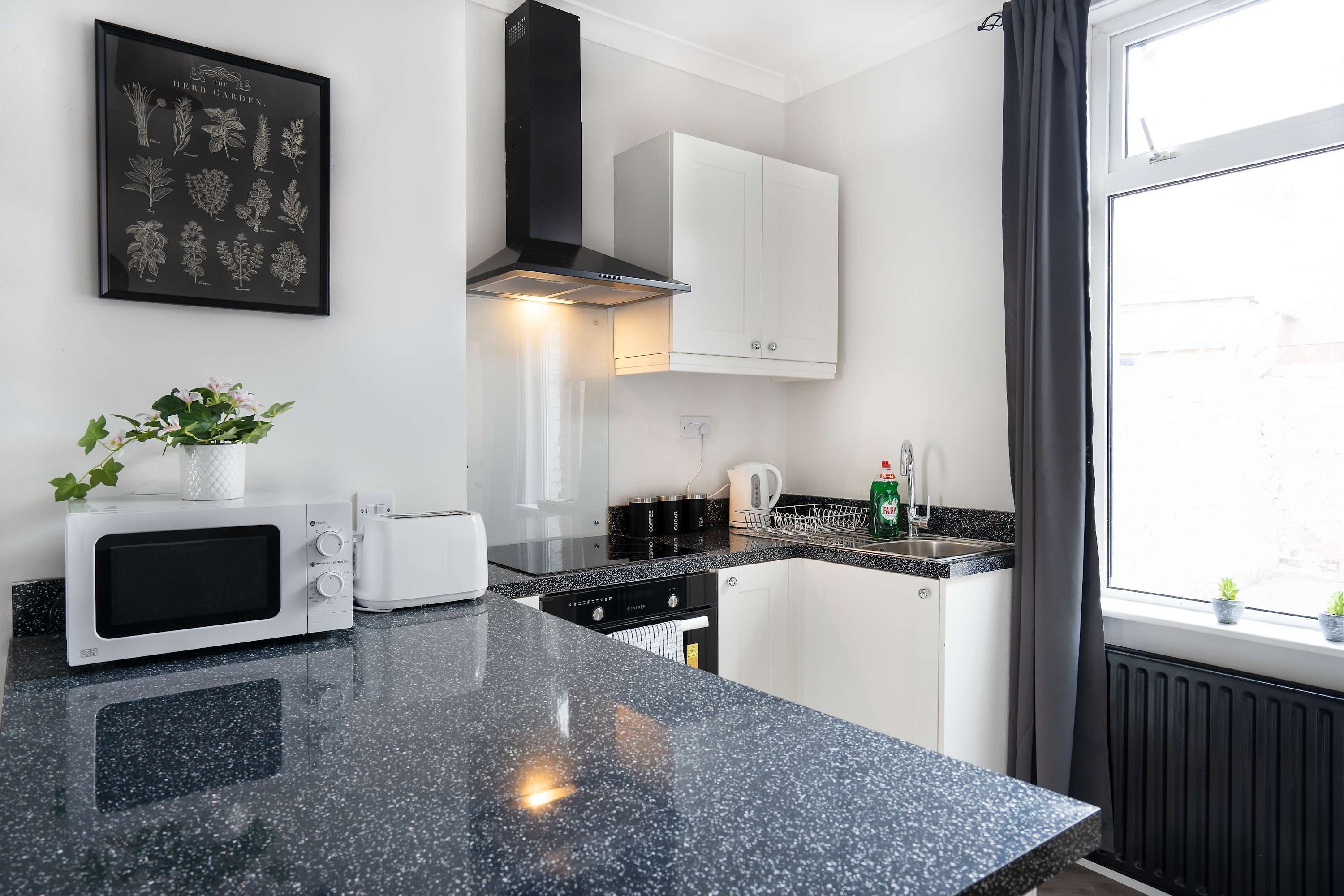 Una cocina moderna con encimera de granito negro, armarios blancos, microondas, tostadora y ventana con cortinas.