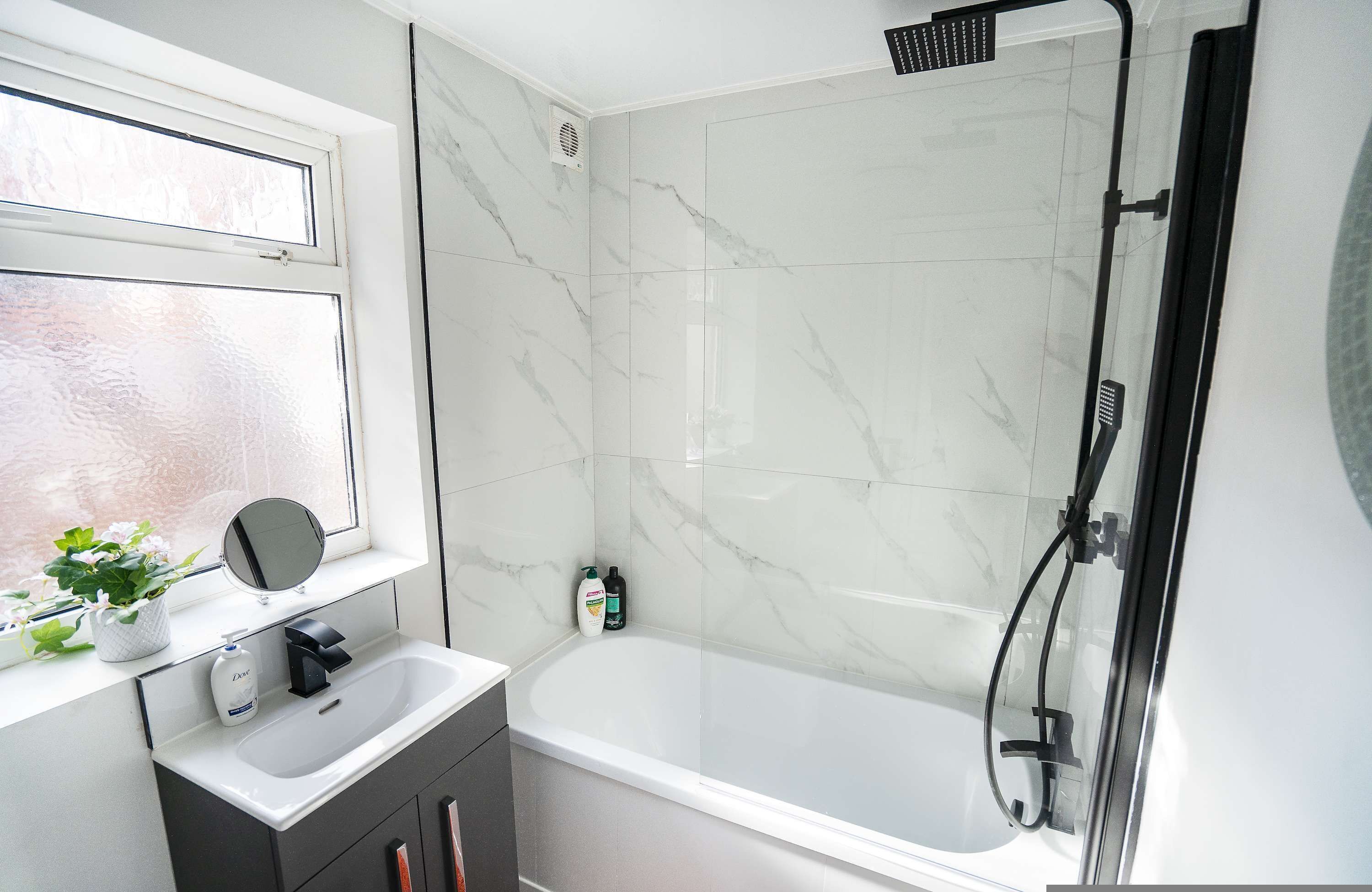 A bathroom with a white bathtub, a shower, a window with a plant, and a mirror.