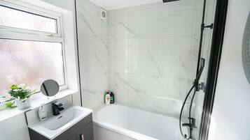 A bathroom with a white bathtub, a shower, a window with a plant, and a mirror.