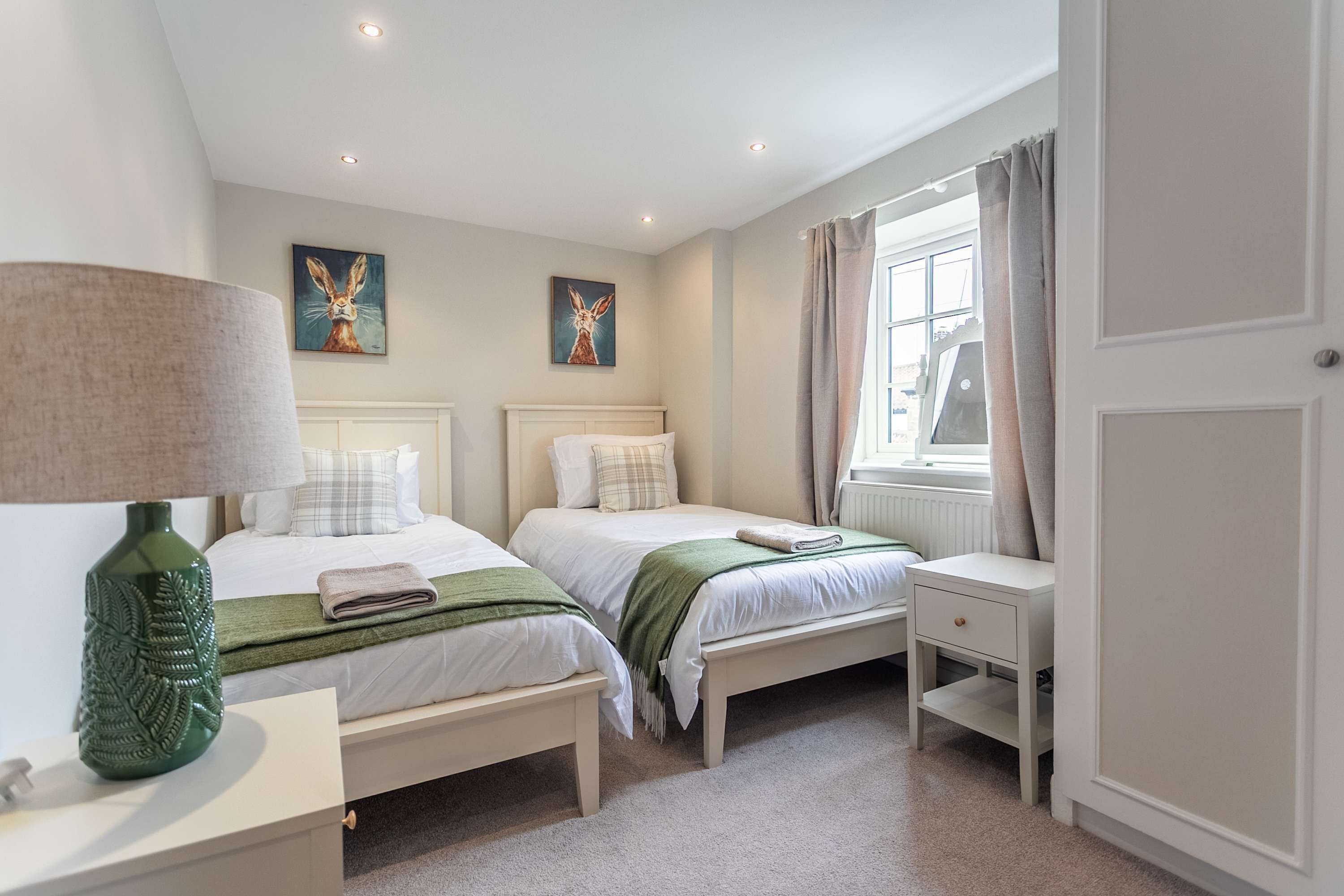 A bedroom with two beds, a bedside table with a lamp, a window with curtains, and artwork on the wall.
