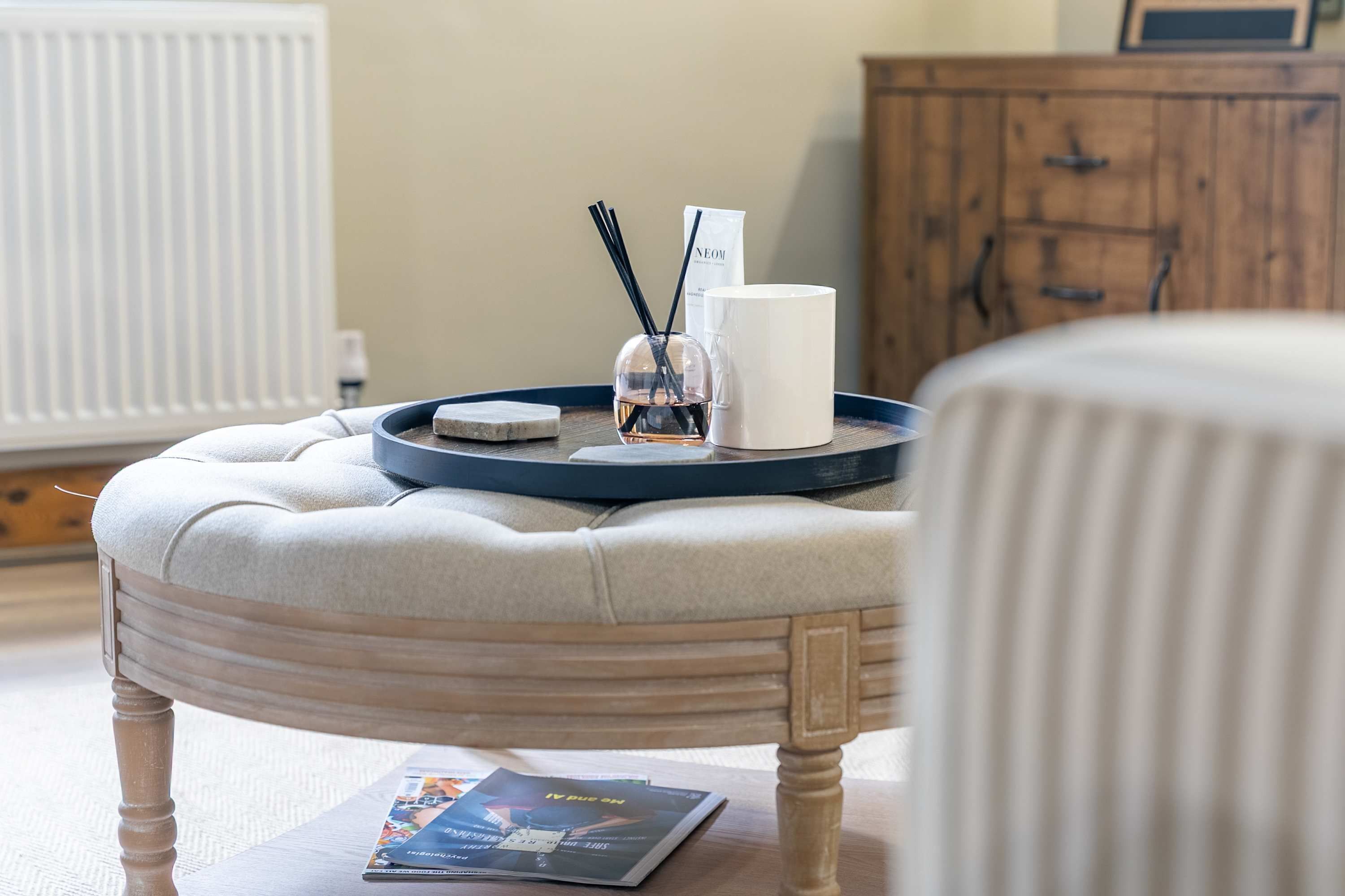 A room with a round, tufted ottoman, a tray with a candle, a glass jar, and a white mug.
