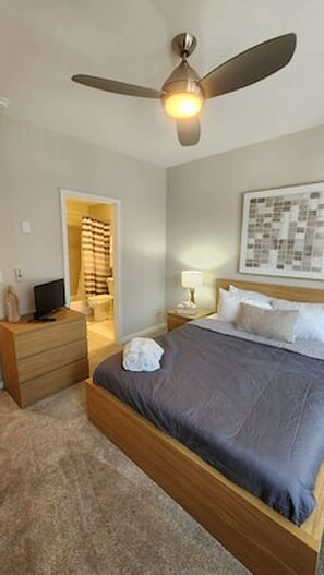 Une chambre à coucher comprenant un lit, une table de chevet, une lampe, un ventilateur de plafond et une porte donnant sur une salle de bain.