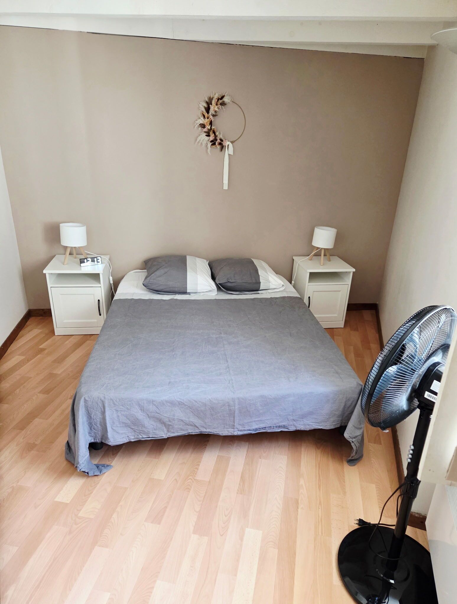 A bedroom with a bed, two bedside tables, a wall-mounted fan, and a decorative wreath.