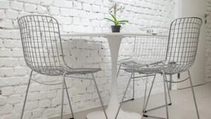 Una pared de ladrillo blanco, una mesa redonda blanca con una planta en maceta, y dos sillas de alambre.