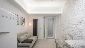 Un espacio habitable moderno y compacto con un sofá blanco, una mesa de comedor con sillas de metal, un radiador y una puerta que da a otra habitación.