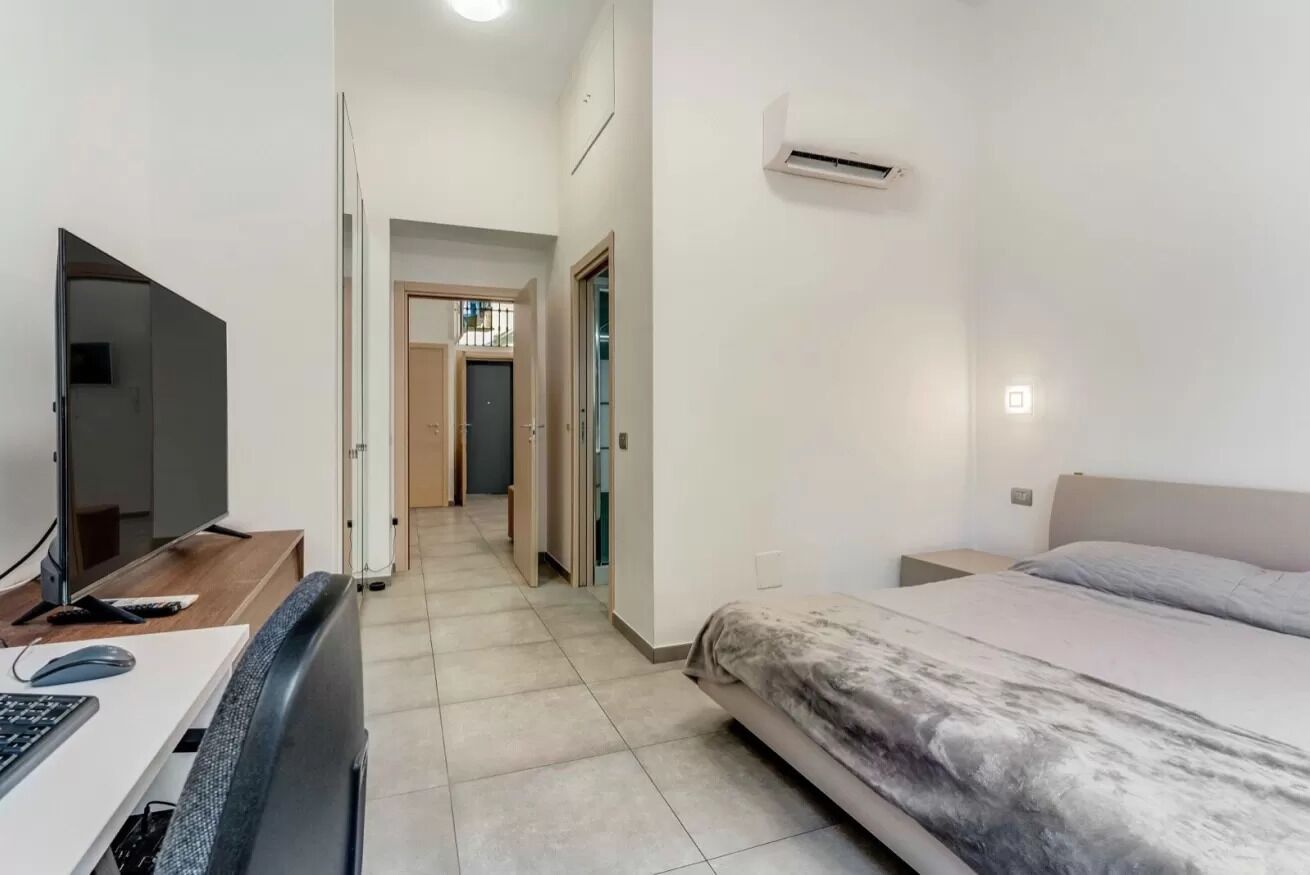 A bedroom with a bed, a desk with a computer, a TV, and an air conditioning unit.