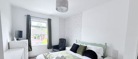 A bedroom with a bed, a desk, a chair, a TV, and a window with curtains.