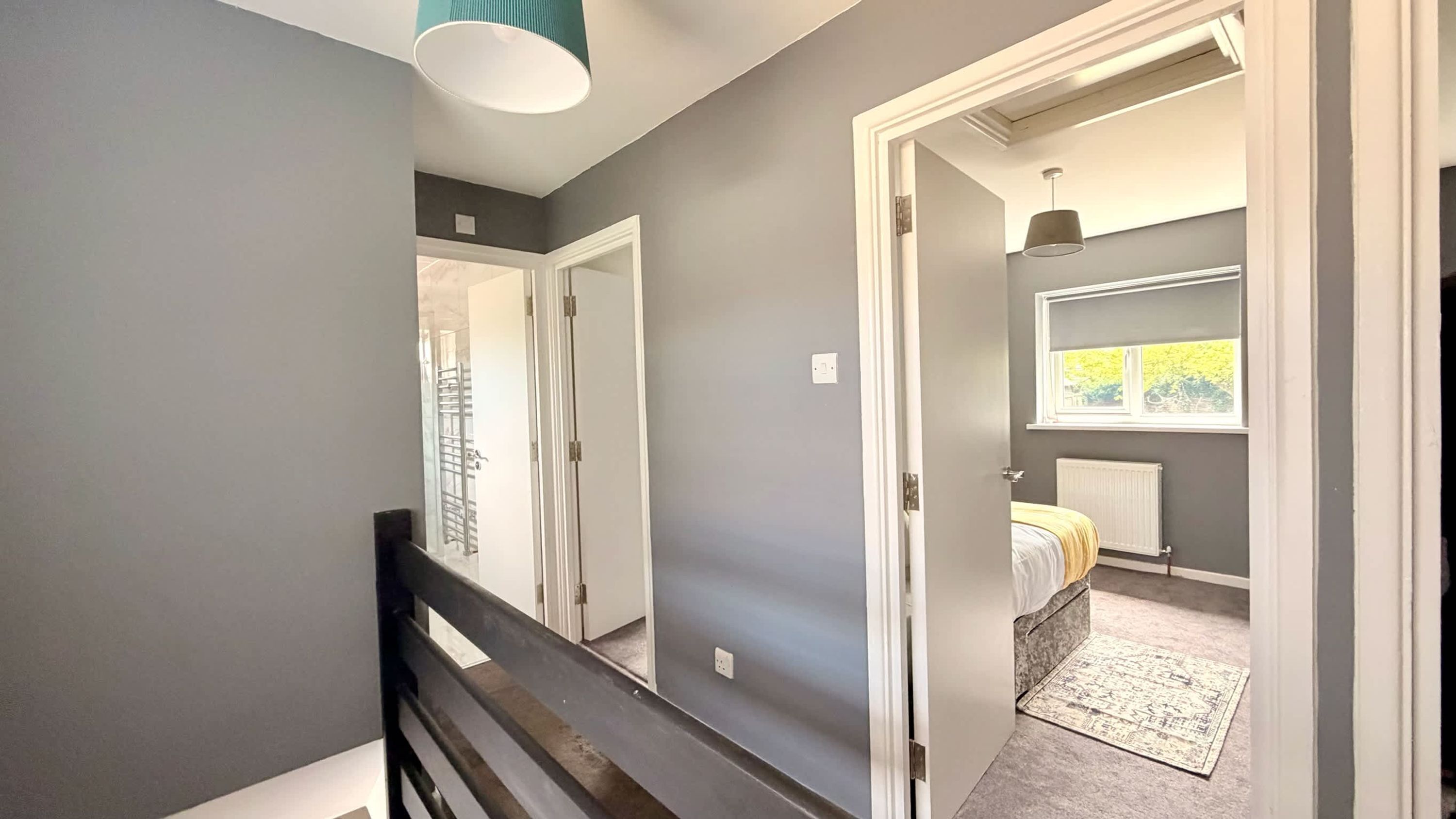 A hallway with a wooden railing, a door leading to a bedroom with a bed and a radiator, and a window with a view of greenery.