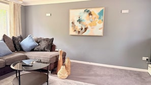 A living room with a grey sofa, patterned cushions, a glass coffee table, a wicker basket, and a painting on the wall.