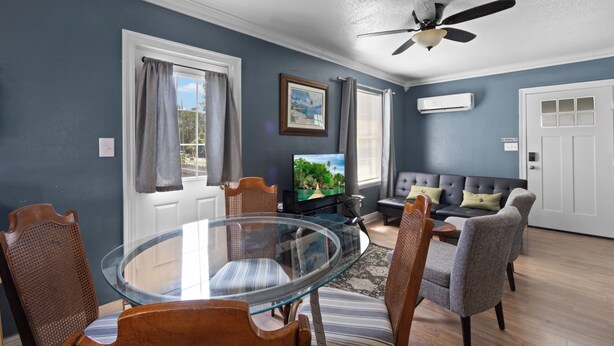 A living room with a glass table, chairs, a sofa, a television, and a ceiling fan. - Welcome to The Blue Bungalow! This cozy 2BR/1BA home is the perfect base for a relaxing Emerald Coast getaway, with a full kitchen, in-unit laundry, WiFi, and a quiet backyard just a short drive from Navarre Beach. (Navarre)