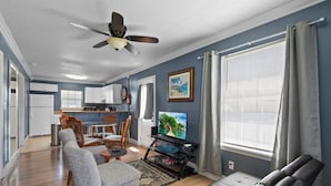A living room with a sofa, a TV on a stand, a ceiling fan, and a dining area with chairs and a table. - Welcome to The Blue Bungalow! This cozy 2BR/1BA home is the perfect base for a relaxing Emerald Coast getaway, with a full kitchen, in-unit laundry, WiFi, and a quiet backyard just a short drive from Navarre Beach. (Navarre)