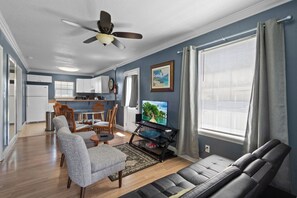 Ein Wohnzimmer mit einer Couch, einem Fernseher auf einem Standfuß, einem Deckenventilator und einem Essbereich mit Stühlen und einem Tisch.