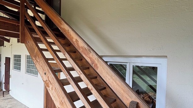 Una escalera de madera que sube hacia un ventanal del segundo piso, con una mesa y sillas abajo.