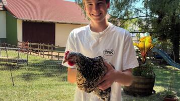 Eine Person hält ein Huhn im Freien, im Hintergrund ein Bauernhaus und ein Zaun.