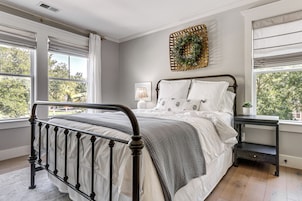 Un dormitorio con cama, mesita de noche, un adorno en la pared y ventanas amplias.