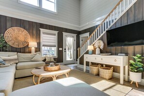 A living room with a sofa, a round wicker table, a TV, a staircase, and a large window. - Walk to Old Town Bluffton! Family Home w/ Balcony (Bluffton)