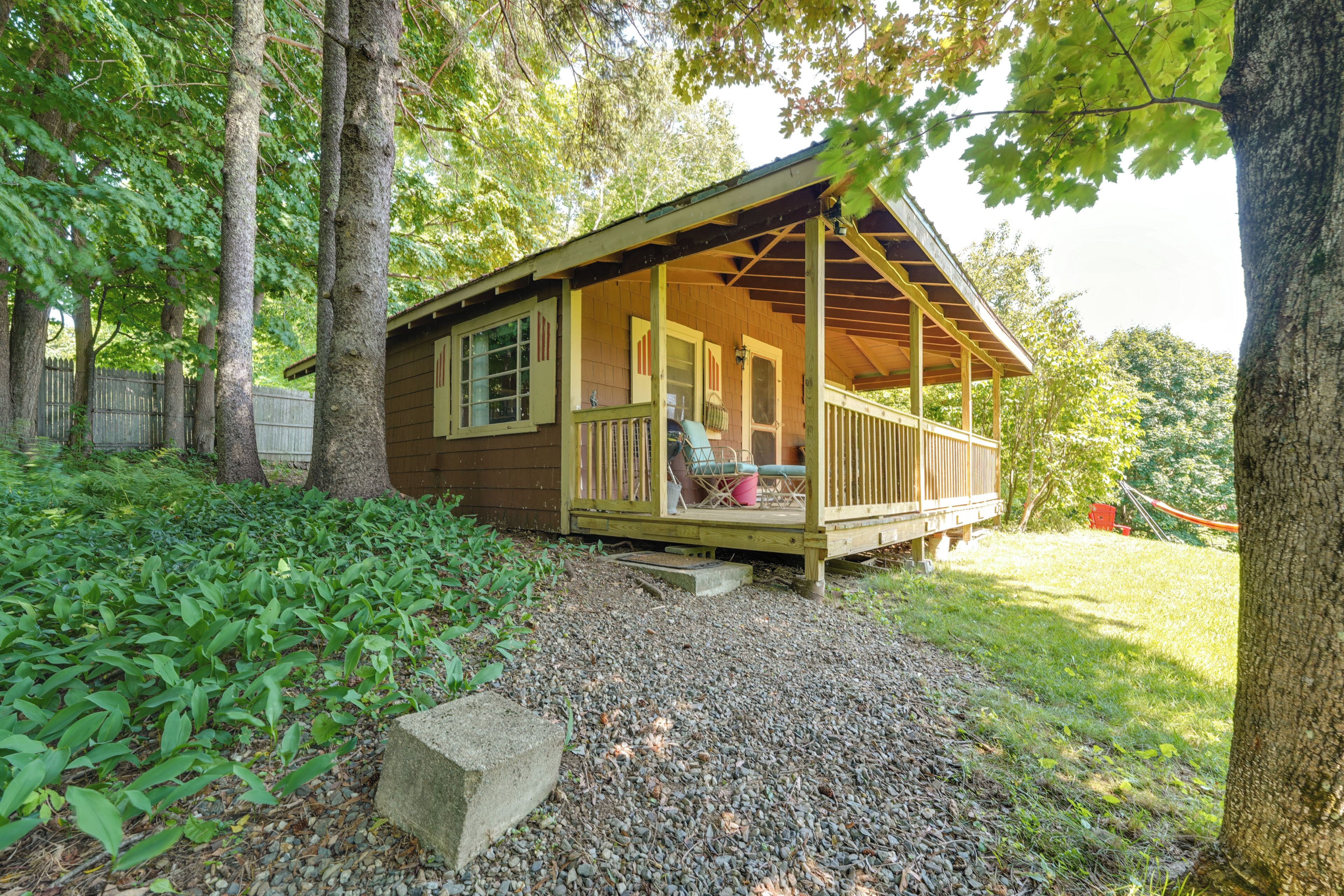 A small wooden cabin with a porch, surrounded by trees and a gravel path.