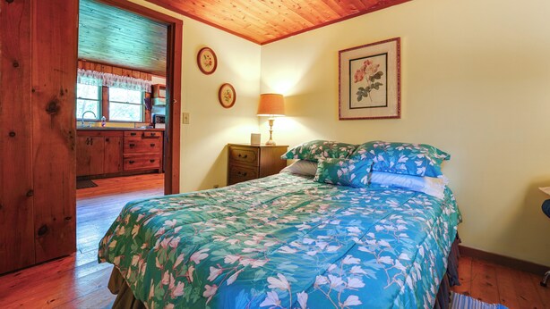 A bedroom with a wooden ceiling, a bed with floral bedding, a nightstand with a lamp, and a window with curtains. - 3 Mi to Hiking & Beaches: Rustic Maine Cabin (Stockton Springs)