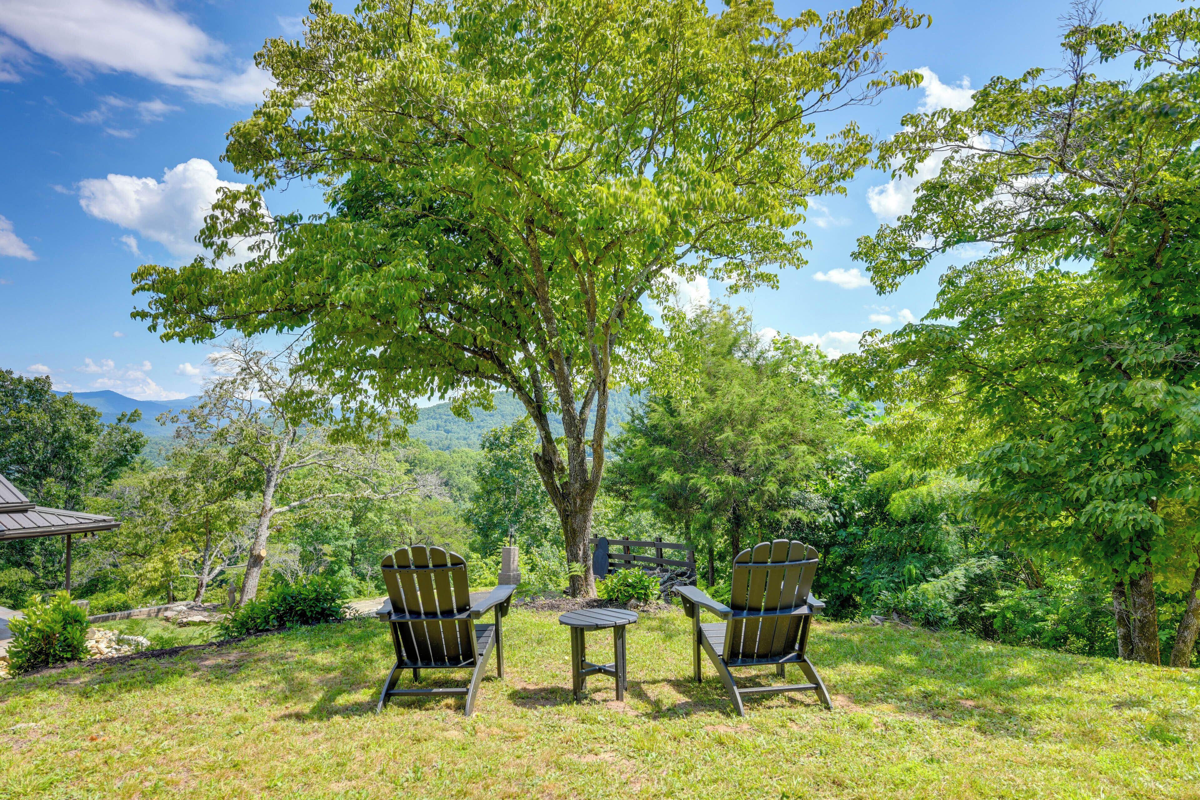 Un espacio exterior tranquilo con dos sillas Adirondack y una mesa pequeña, rodeado de exuberante vegetación y con árboles y montañas lejanas al fondo.