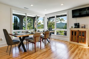 A dining area with a wooden table and chairs, large windows with a view of trees and mountains, and a kitchen area with a coffee machine and cabinets. - 32-acre Escape w/ Trails & Games! Gateway to Gsmnp (Franklin)