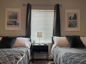 Quarto com duas camas, mesa de cabeceira, luminária e janela com persianas.