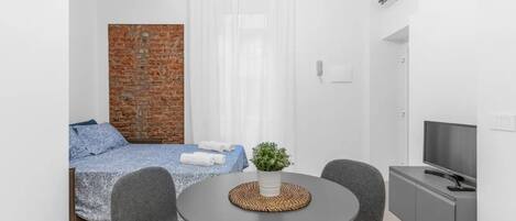 A modern room with a bed, a small table with chairs, a TV, and a brick wall.