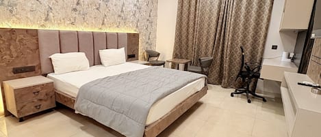 A modern bedroom with a large bed, bedside tables, a desk, a chair, a ceiling fan, and a bathroom with a mirror and sink.