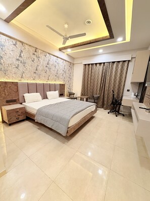 Une chambre moderne avec un grand lit, des tables de chevet, un bureau, une chaise, un ventilateur de plafond et une salle de bain avec un miroir et un lavabo.