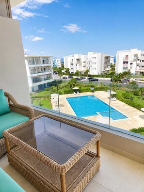 A balcony with a wicker chair and a glass table, overlooking a swimming pool and residential buildings.