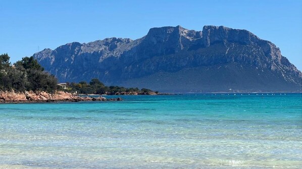 A coastal landscape with clear turquoise waters, a rocky shoreline, and a prominent mountain in the background.